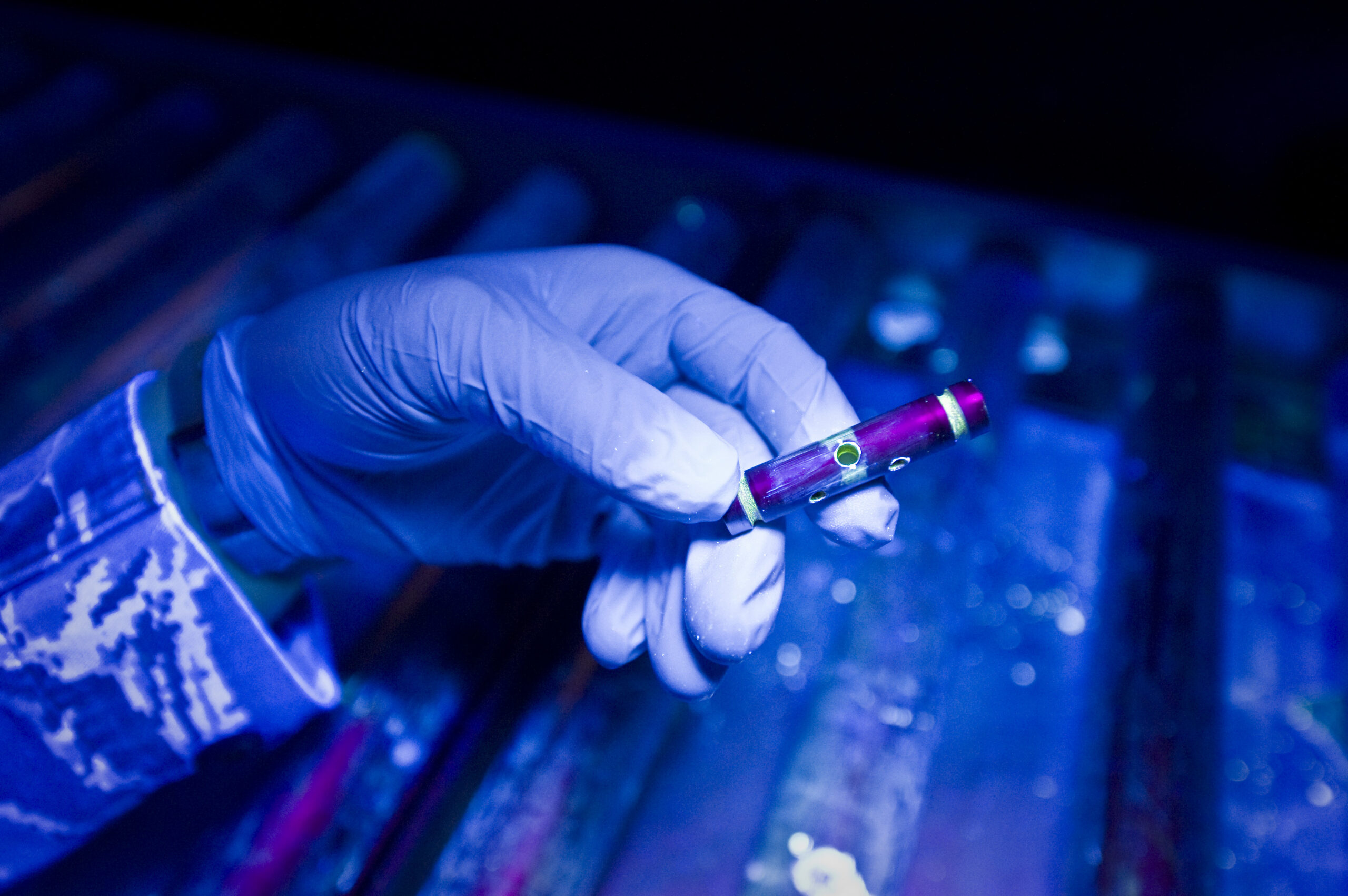 Military NDT technician performing fluorescent penetrant inspection on an aircraft pin under ultraviolet black light, revealing surface cracks invisible to the naked eye — illustrating the precision and expertise behind nondestructive testing methods used across aerospace and defense