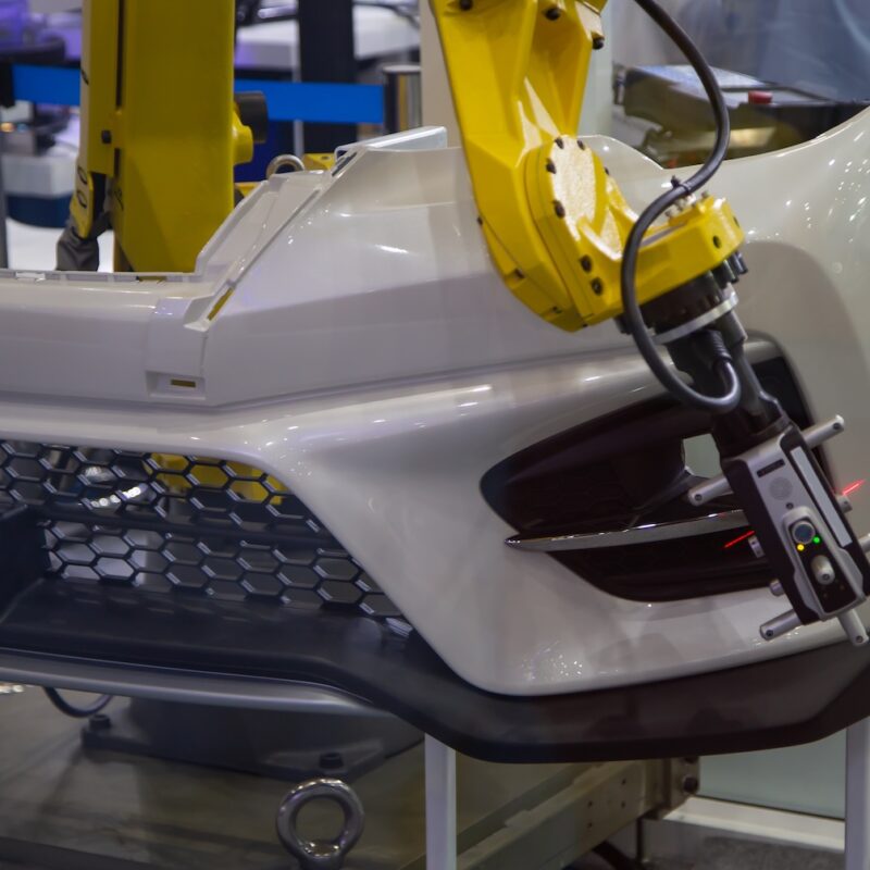 A yellow robotic arm uses a 3D laser scanner for quality control on a composite car bumper, a key process for collecting the physical data needed for composite simulation validation.