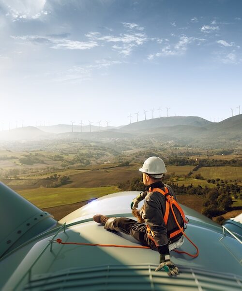 An engineer in safety gear performing maintenance on a wind turbine, with a view of a large wind farm.