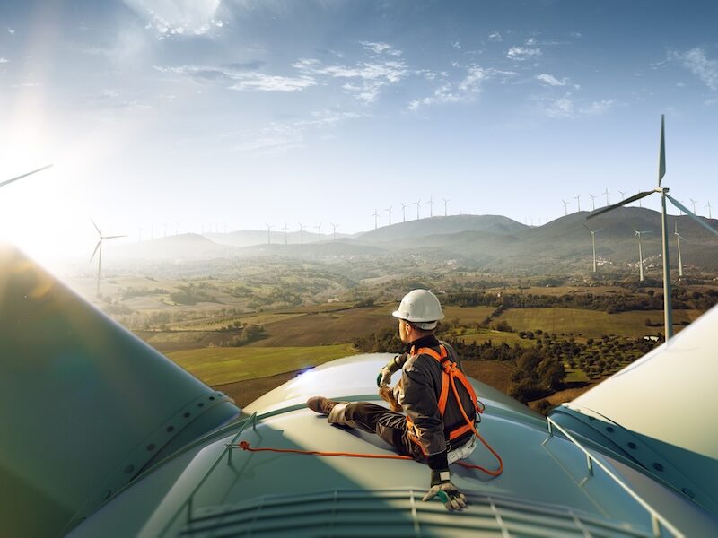 An engineer in safety gear performing maintenance on a wind turbine, with a view of a large wind farm.